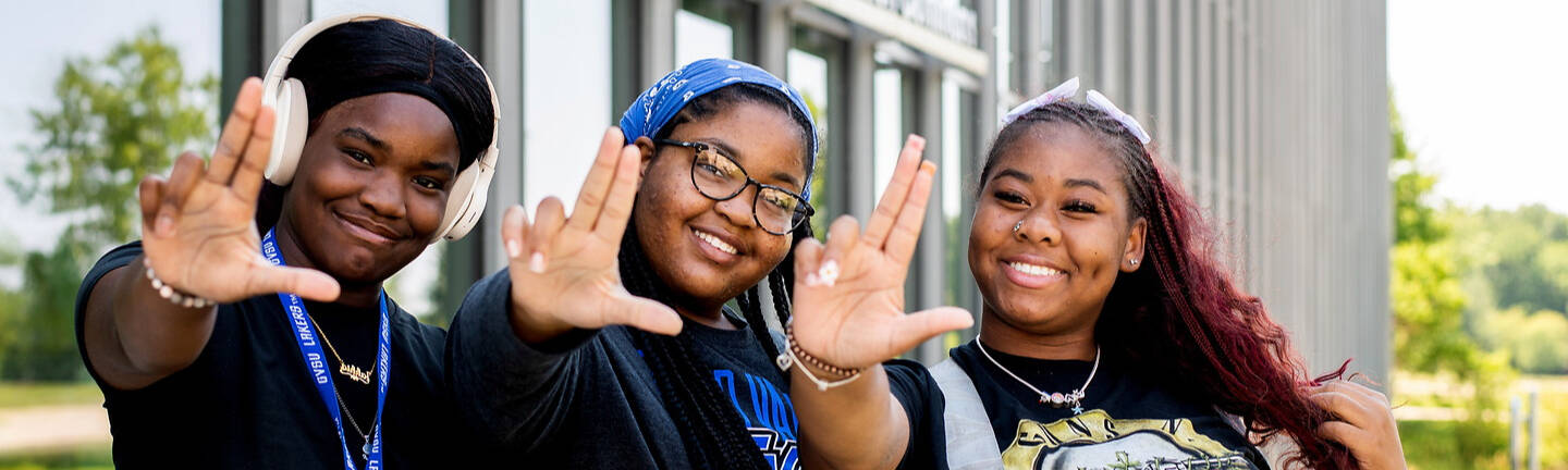 Three young individuals pose cheerfully outside a library building, making hand gestures toward the camera.